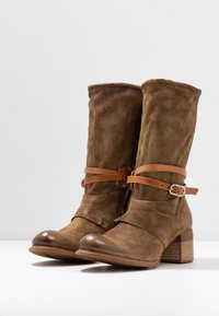 Brown suede ankle boots with a mid-heel, featuring leather straps and a buckle detail, and a rounded toe design. Zipper on the side.