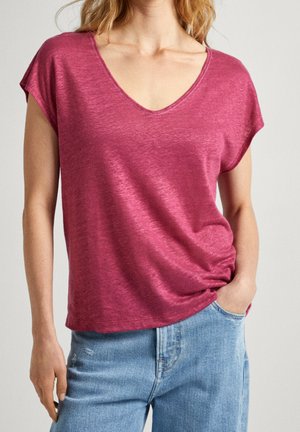 Kobieta w luźnej różowej bluzce z krótkim rękawem i dekoltem w serek, połączonej z jasnoniebieskimi, wysokim stanem dżinsami, z jedną ręką w kieszeni, na jednolitym tle.