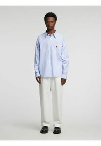 Svetlo modra srajca na gumbe z modrimi črtami in žepom na prsih ter okrasno nalepko, kombinirana z razširjenimi belimi hlačami in črnimi čevlji.