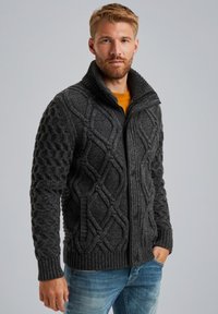 Jersey gris oscuro con cuello alto, diseño de trenzas texturizadas, puños acanalados y cierre de cremallera. Lleva una camisa amarilla debajo.