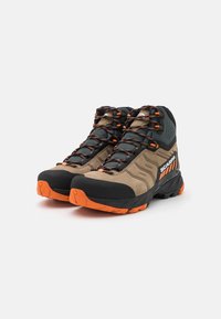 Buty trekkingowe z brązową zamszową górą, czarnymi akcentami, pomarańczową gumową podeszwą i szarymi siateczkowymi detalami. Posiadają wzmocniony nos i teksturowane sznurowadła.