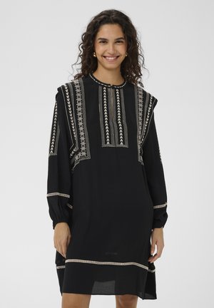 Žena sa usmieva, oblečená v čiernych dlhých šatách s bielou geometrickou výšivkou pozdĺž rukávov a prednej časti, stojaca pred jednoduchým pozadím.