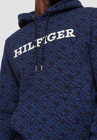 Modrá mikina s kapucí se vzorem, obsahující texturovanou látku, velké bílé logo "HILFIGER" a nastavitelné šňůrky na kapuci.