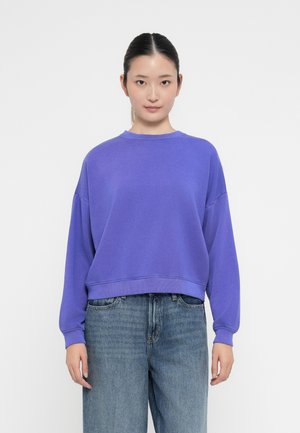 Jonge vrouw met zwart haar dat vastgebonden is, gekleed in een losse paarse sweater en blauwe spijkerbroek, staande voor een effen witte achtergrond.
