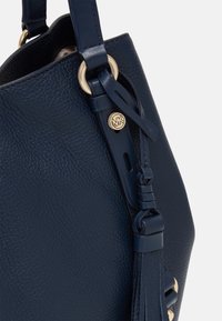 Námořnická kožená taška ve tvaru kbelíku s texturovanou povrchovou úpravou, zlatými detaily a akcentem v podobě uvázané šňůrky. Obsahuje viditelné logo značky.