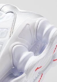 Ténis desportivo branco com uma parte superior em tecido texturado, detalhes em plástico liso e uma sola em camadas distinta com detalhes vermelhos.