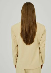 Beige přizpůsobený blazer s dlouhými rukávy, vyrobený z hladké látky, s přímým střihem a rozparkem na zadní straně. Viditelné dlouhé rovné vlasy.