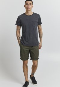 T-shirt en coton gris avec une poche sur la poitrine, associé à un short olive roulé. Des baskets noires complètent la tenue décontractée.