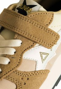 Dettaglio di una sneaker beige in crosta con strap in velcro, lacci bianchi e logo a stella nera con testo dorato "SUN68" sulla linguetta.