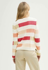 Femme aux longs cheveux blonds portant un pull en crochet multicolore à rayures horizontales rouges, pêche, beige et blanches, associé à un pantalon beige.