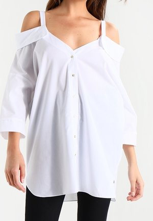 Kvinde iført løs hvid cold-shoulder skjorte med knapper, 3/4 ærmer og tynde skulderstropper, kombineret med sorte bukser.