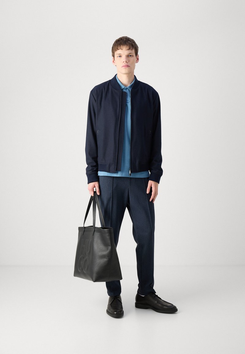 Granatowa kurtka bombowca na lekko niebieskim polo, zestawiona z ciemnymi, teksturowanymi spodniami. Czarna torba na ramię z wytłoczonym logo oraz czarne buty.