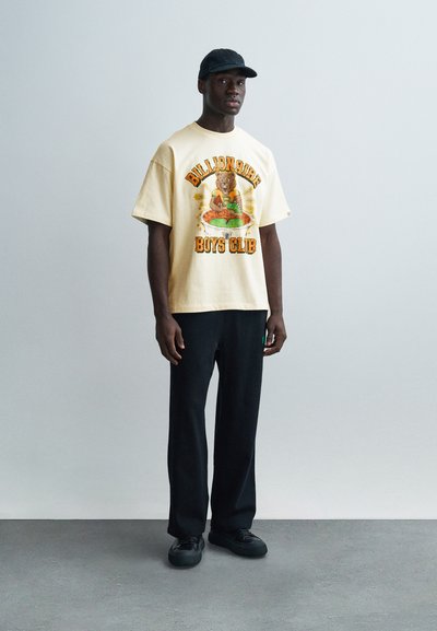 Κοντό μπεζ μπλουζάκι με γραφικό εκτύπωμα που απεικονίζει μια αρκούδα και το κείμενο «Billionaire Boys Club», σε συνδυασμό με μαύρο παντελόνι και μαύρα παπούτσια.