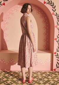 Rožnata in rumena cvetlična obleka z oprijetim zgornjim delom in razširjenim krilom, kombinirana s rdečimi čevlji. V ozadju je rožnat lok z dekorativnimi vzorci.