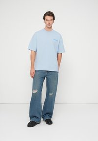 Φαρδιά γαλάζια βαμβακερή μπλούζα με στρογγυλή λαιμόκοψη, κοντά μανίκια και μικρό λογότυπο στο στήθος, συνδυασμένη με τζιν παντελόνι με σκισίματα.