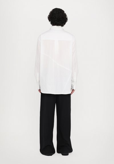 Camisa blanca de manga larga con cuello, que presenta un panel trasero transparente y puños abotonados, combinada con unos pantalones negros holgados.