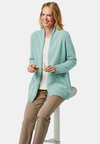 Helles blaues Strickjäckchen aus weichem Material, mit einem Schalkragen, zwei Vorderseiten-Taschen und gerippten Ärmeln, getragen über einem weißen Oberteil und beigen Hosen.