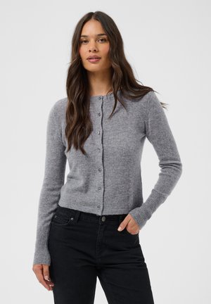 Kvinde med langt brunt hår iført en grå cardigan med knapper og sorte bukser, står med den ene hånd i lommen mod en ensfarvet baggrund.