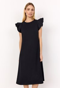 Frau, die ein schwarzes Midikleid mit gerafften kurzen Ärmeln trägt, steht vor einem schlichten weißen Hintergrund.