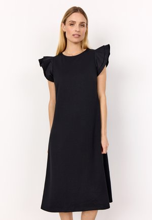 Frau, die ein schwarzes Midikleid mit gerafften kurzen Ärmeln trägt, steht vor einem schlichten weißen Hintergrund.