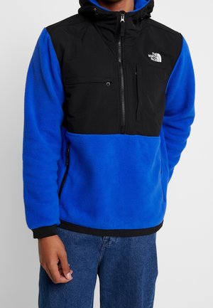 Man draagt een blauwe en zwarte fleece trui met ritszakken op de borst en zijkanten, met het logo van The North Face op de borst.