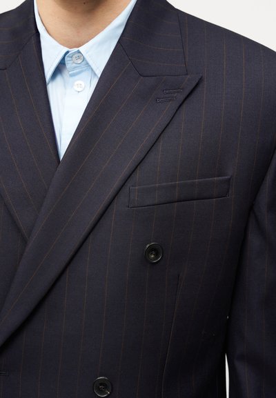 Σκούρο μπλε μπλέιζερ με διπλό πέτο και ρίγες, με καφέ ρίγες, πέτο τύπου notch και μαύρα κουμπιά. Κάτω φαίνεται ένα ανοιχτό γαλάζιο πουκάμισο.