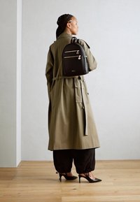 Abrigo trench color caqui con cintura ceñida, acompañado de pantalones negros holgados y zapatos de tacón negro. Se lleva una pequeña mochila negra en la espalda.