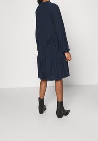 Robe bleu marine à volants avec des manches longues, en tissu texturé et avec des motifs subtils. Portée avec des bottines noires à la cheville. Vue de derrière.