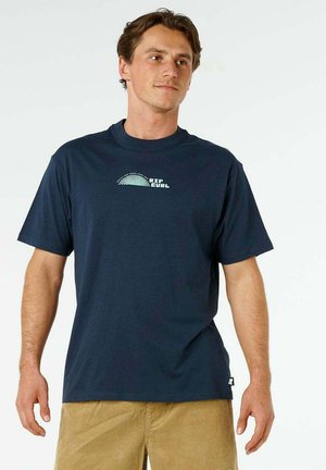 Mężczyzna ubrany w granatową koszulkę Rip Curl i jasnobrązowe spodnie, stoi w zrelaksowanej postawie na jednolitym białym tle.