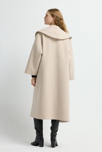 Donna con capelli ondulati che indossa un lungo cappotto beige con colletto ampio e stivali neri alti fino al ginocchio con tacco, che si trova di lato contro uno sfondo bianco.