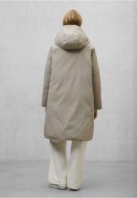 Abrigo acolchado largo beige claro con una capucha grande, textura suave, diseño minimalista y cortes laterales. Combinado con pantalones blancos de campana y zapatillas.