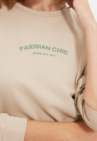 Camicia beige a maniche lunghe con testo verde "PARISIAN CHIC" e "POSE ALL DAY", indossata da una persona con le braccia incrociate e le maniche arrotolate.