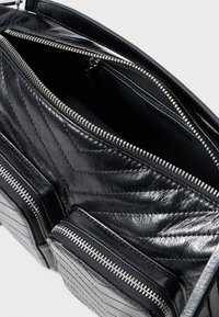 Saco de couro preto com costura em ziguezague, zíperes metálicos, bolso frontal e compartimento principal aberto sobre fundo branco.