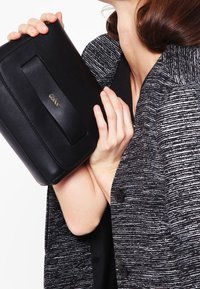 Musta nahkakotelo, jossa on etuflap ja nauha-akcentti. Sileä pinta ja kompakti muoto. Yhdistetty harmaaseen teksturoituun takkiin.