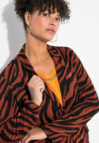 Blazer a strisce zebra nere e arancioni con tessuto testurizzato, dotato di colletto e maniche lunghe. Indossato su un top arancione brillante con scollatura gioiello.
