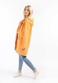Imperméable orange avec capuche, comportant des boutons-pression et deux poches avant. Associé à un jean bleu clair et des baskets blanches.
