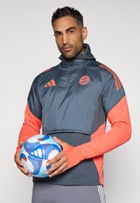 Grå og koral Adidas jakke med halvlynlås, der har FC Bayern Münchens logo og tre striber som accent. Holder en multicolor fodbold.