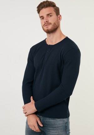 Dunkelblaue Langarm-Henley-Shirt aus strukturiertem Stoff mit drei Knöpfen am Ausschnitt, kombiniert mit hellblauen Jeans.
