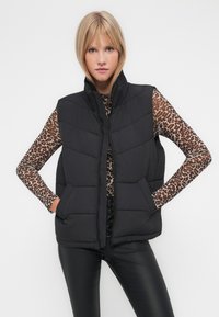 Zwarte gewatteerde vest met een hoge kraag, voorzien van een ritssluiting en zijzakken. Model draagt een lange mouwen top met luipaardprint eronder.