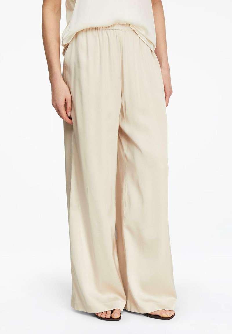 Frau trägt locker sitzende, beige, weit geschnittene Hosen mit elastischem Bund, kombiniert mit schwarzen, offenen Sandalen.