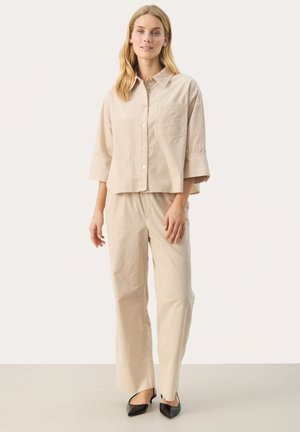 Beige, cropped knoopshirt met een borstzak; gecombineerd met losse beige broeken. Zwart, puntige schoenen maken de outfit compleet.