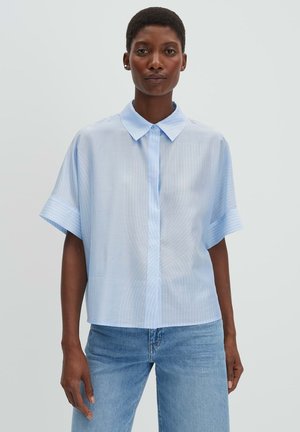 Frau trägt ein hellblaues kurzärmeliges Hemd mit Kragen und hellblaue Jeans, steht vor einem schlichten Hintergrund.