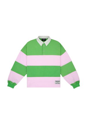 Man wearing oversized green and pink striped rugby shirt with white collar and dark gray pants, standing against plain background.