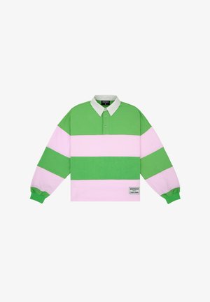 Mężczyzna w za dużej zielono-różowej prążkowanej koszulce rugby z białym kołnierzem i ciemnoszarych spodniach, stojący na jednolitym tle.