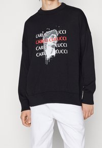 Schwarzer Sweatshirt mit lockerer Passform, ausgestattet mit einem grafischen Druck eines Statuen-Kopfes und dem Markennamen in Rot und Weiß. Weicher Stoff, gerippte Bündchen.