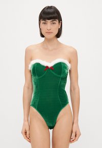 Bodysuit sans manches en velours vert avec des bordures en fausse fourrure blanche et un petit nœud rouge au centre de la poitrine. Ouvertures de jambes hautes.