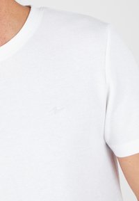 Camiseta blanca de manga corta en tejido texturizado. Presenta un cuello redondo y un sutil logo en el pecho. Diseño limpio con detalles mínimos.