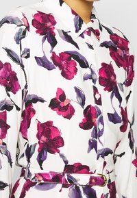 Camicia bianca con stampa floreale grande in magenta e viola, dotata di una cintura floreale coordinata con fibbia dorata.