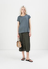 Grå kortærmet top parret med en lang, mørkegrøn knap-front nederdel. Tilbehøret inkluderer en lille beige håndtaske og sorte flip-flops.
