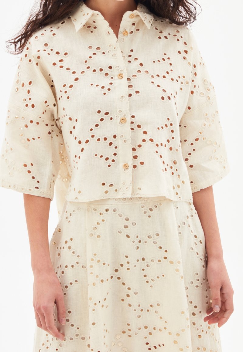 Femme portant un chemisier crème boutonné et une jupe avec des motifs découpés circulaires et des manches 3/4.